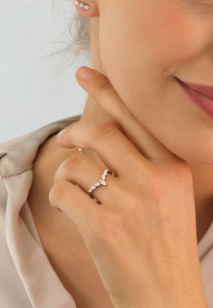 Glänzender silberner Ring mit einem V-förmigen Design, besetzt mit marquiseförmigen Steinen. Glatte Textur; wird am Mittelfinger getragen und ergänzt die Ohrstecker.