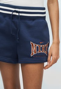 Σκούρο μπλε αθλητικό σορτς Nike με λευκή ριγέ ζώνη μέσης και κορδόνι, με πορτοκαλί λογότυπο «NIKE» στο αριστερό μηρό.