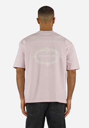 Rövid hajú férfi világos rózsaszín pólóban, amelyen díszes fehér felirat olvasható: "Moments to Memories", és fekete nadrágot visel, háttal áll.