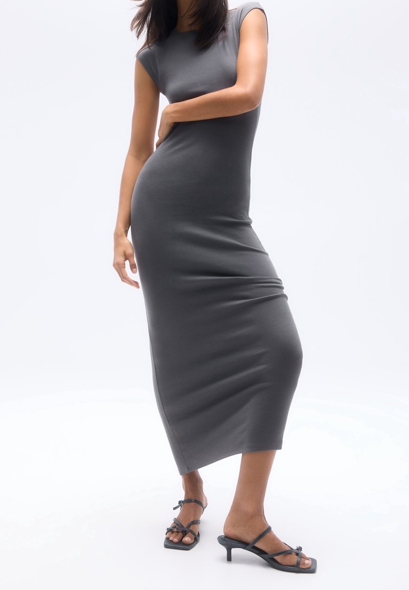 Femme portant une robe midi grise ajustée sans manches et des sandales noires à brides avec petit talon sur fond blanc.