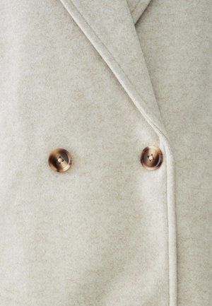 Divkrāsu mētelis gaiši bēšā vilnā, ar diviem brūniem, apaļiem pogām un gludu, teksturētu audumu ar smalku rakstu.