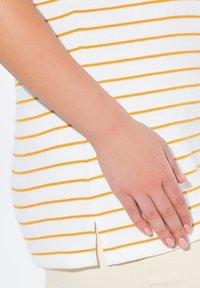Bílý top se žlutými vodorovnými pruhy, s bočním rozparkem a texturovaným bavlněným materiálem. Ruka spočívá na spodním lemu.