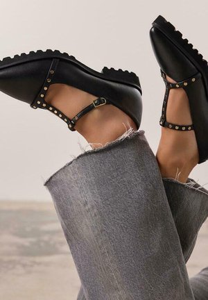 Zapatos de cuero negros con una tira en forma de T, tachuelas doradas que acentúan la tira, suela de goma y punta redonda, combinados con unos jeans de mezclilla gris desgastados.