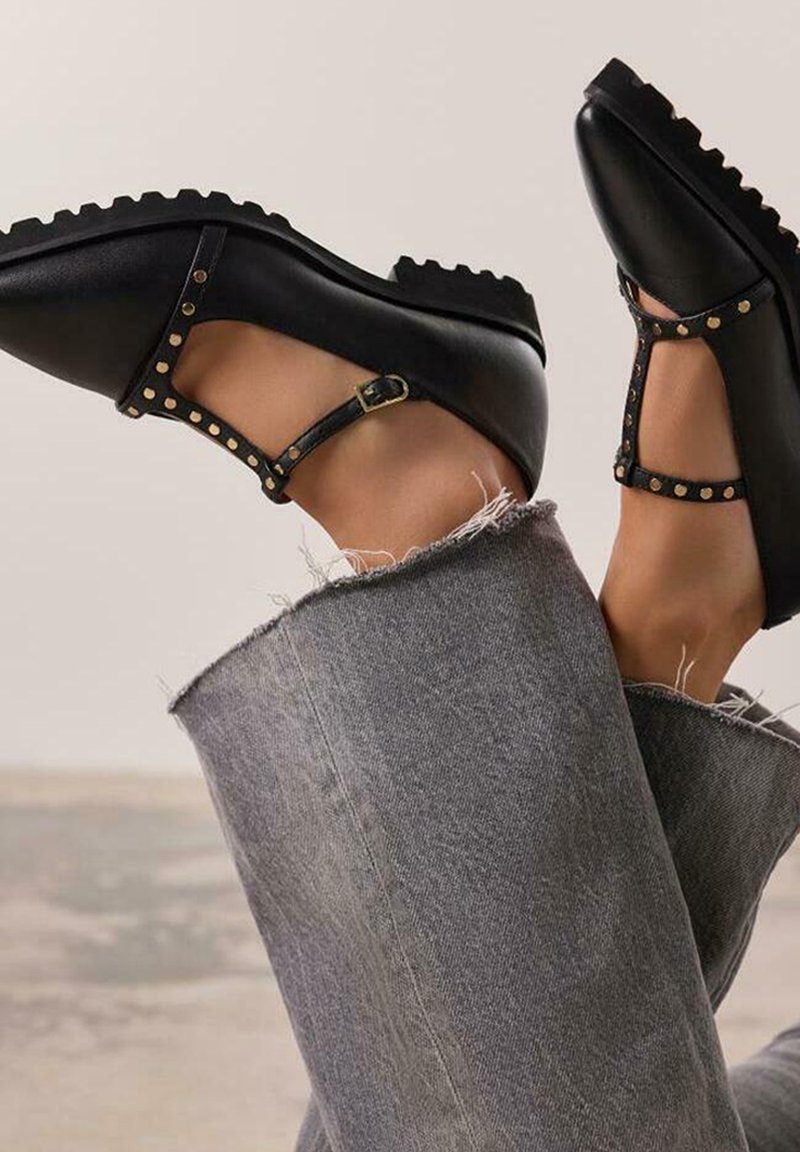 Chaussures en cuir noir avec une lanière en T, des clous dorés accentuant la lanière, une semelle en caoutchouc et un bout arrondi, associées à un jean gris usé.