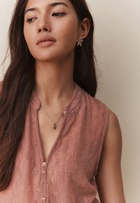 Top senza maniche con texture pesca, scollo a V e chiusura con bottoni, abbinato a un orecchino a forma di stella marina dorata e a una delicata collana con un ciondolo a forma di lettera.