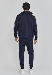 Marine trainingspak van zacht materiaal, met een opstaande kraag en contrasterende gouden accentstrepen langs de mouwen en broeken.