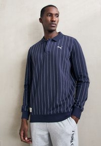 Mørkeblå langærmet polo med hvide lodrette striber, der har krave og logo på brystet; parret med grå sweatpants.