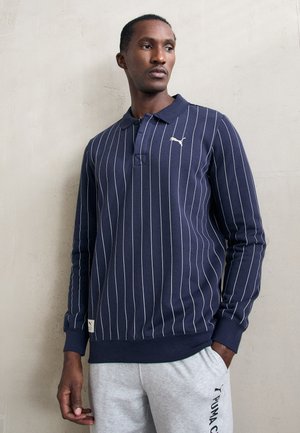 Navy blue long-sleeve polo with white vertical stripes, featuring a collar and logo on the chest; paired with gray sweatpants.