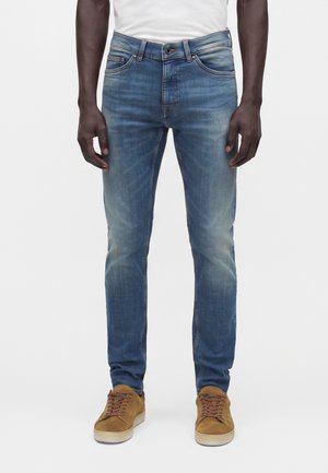 Homme portant un jean slim bleu avec des détails délavés et des baskets en daim marron, debout devant un fond blanc.