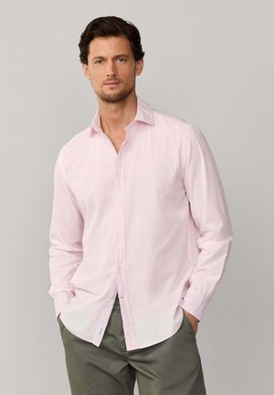 Homme aux cheveux courts et foncés portant une chemise rose pâle boutonnée et un pantalon vert olive, debout les mains dans les poches devant un fond uni.