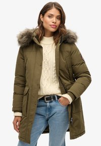 Parka de color verde oliva con capucha forrada de piel desmontable, con cierres a presión, dos bolsillos frontales y un interior cálido. Suéter blanco de punto de cable debajo.