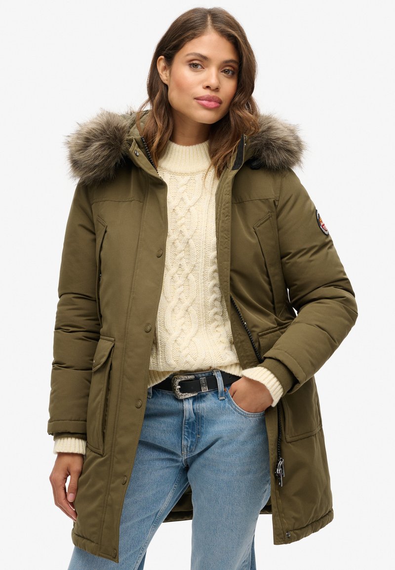 Parka de color verde oliva con capucha forrada de piel desmontable, con cierres a presión, dos bolsillos frontales y un interior cálido. Suéter blanco de punto de cable debajo.