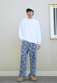 Mladý muž stojící proti béžové zdi, oblečený v bílé košili s dlouhým rukávem, vzorovaných modrých džínách, hnědých botách a bílé čepici.