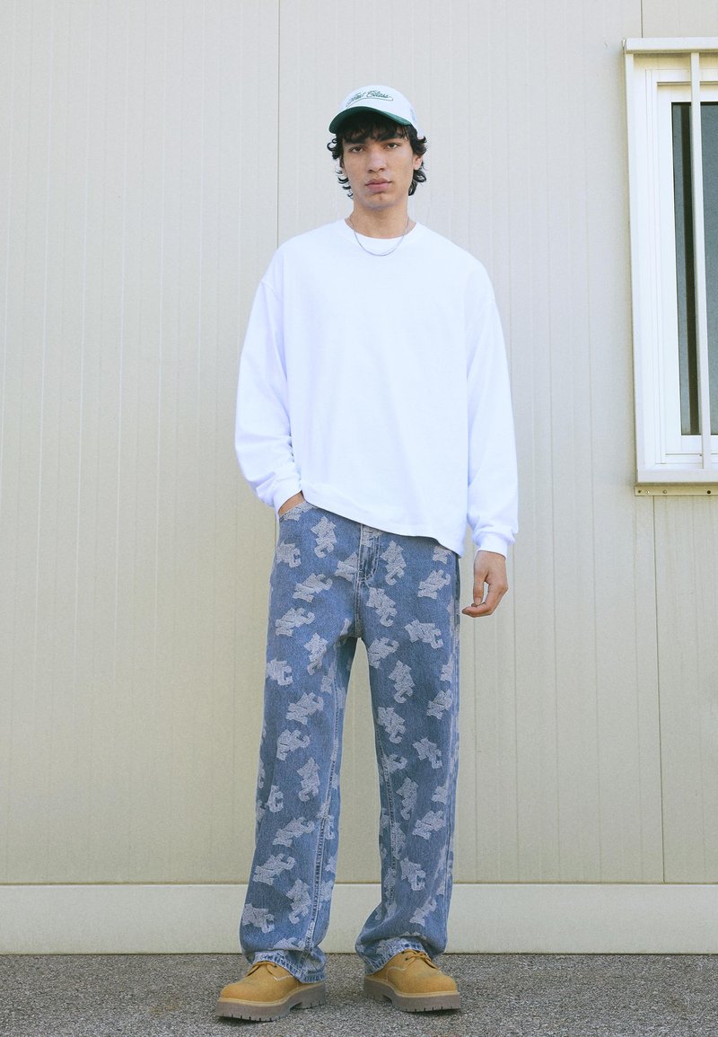 Mladý muž stojící proti béžové zdi, oblečený v bílé košili s dlouhým rukávem, vzorovaných modrých džínách, hnědých botách a bílé čepici.