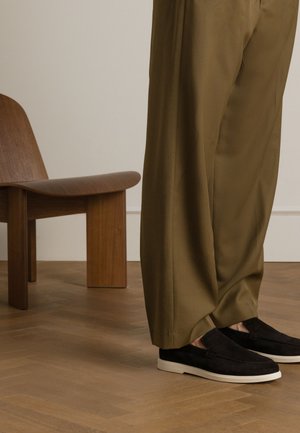 Zwarte suède instapschoenen met een witte rubberen zool, gecombineerd met olijfgroene loszittende broek, tegen een achtergrond van een houten stoel.