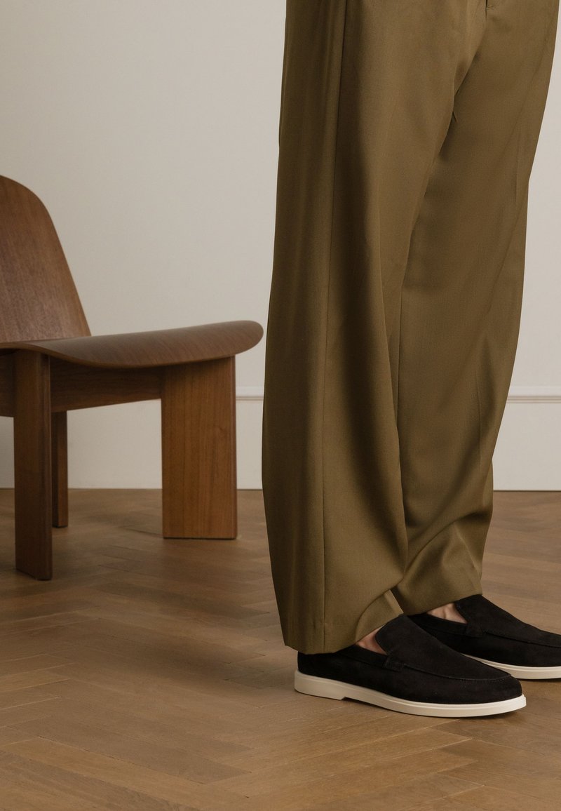 Black suede slip-on shoes with a white rubber sole, paired with olive green loose-fitting pants, set against a wooden chair backdrop.