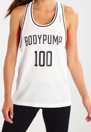 Kobieta nosząca biały sportowy top na ramiączkach z czarną lamówką z napisem "BODYPUMP 100" i czarne legginsy.
