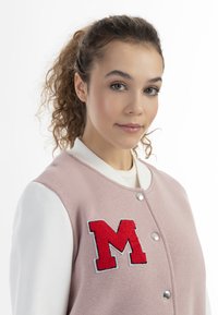 Rosa Varsity-Jacke mit weißen Ärmeln, die mit einem großen roten Buchstaben "M" Aufnäher und silbernen Druckknöpfen versehen ist. Weicher Stoff und lässiges Design.
