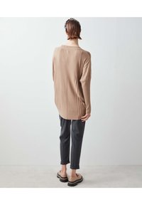 Pull en maille beige à la coupe décontractée, avec un élément de design diagonal, associé à un pantalon gris ample et des chaussures de couleur claire.