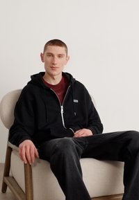 Černá mikina na zip s logem, nošená přes hnědou košili. Osoba je usazena na světlém křesle, na sobě má tmavé džíny a krátké vlasy.