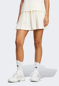 Jupe de tennis plissée crème au design fluide, associ�ée à des chaussures de sport blanches et des chaussettes arborant le logo Adidas.