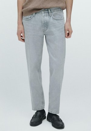 Uomo che indossa jeans grigi chiaro a gamba dritta, mocassini neri in pelle e un maglione beige lavorato a maglia su sfondo bianco semplice.