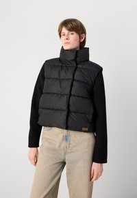 Colete puffer preto cropped com colarinho alto, apresentando secções acolchoadas. Usado sobre um suéter preto, combinado com jeans de tom claro.