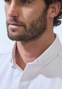 Weißes Hemd mit strukturierter Stoffoberfläche, ausgestattet mit einer Knopfleiste und sichtbaren Nähten. Der Kragen des Hemdes ist halbsteif.