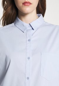 Woman wearing light blue buttoned shirt with pointed collar and chest pocket, shown from shoulders to chin.