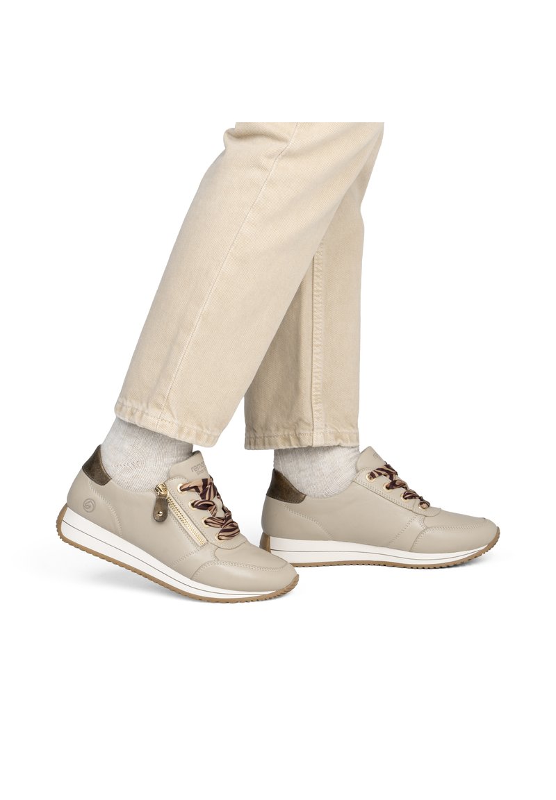 Baskets beiges avec des lacets marron et une fermeture éclair sur le côté, portées avec un pantalon beige clair et des chaussettes blanches, sur fond blanc.