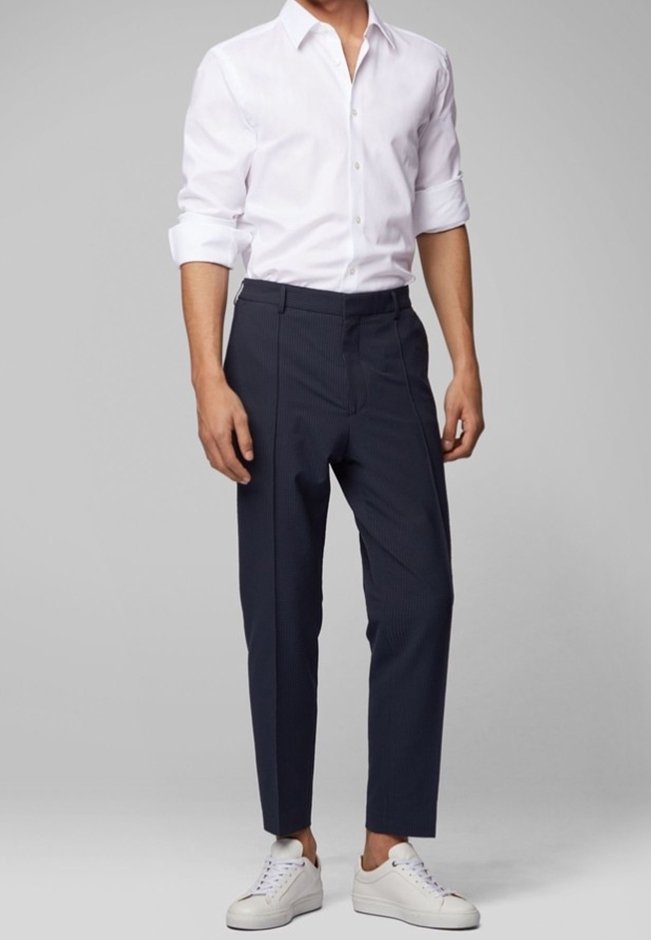 Homme portant une chemise blanche retroussée, un pantalon noir sur mesure et des baskets blanches, se tenant contre un fond gris uni.