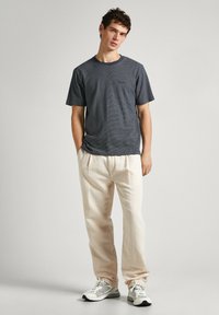 Camiseta a rayas azul oscuro con cuello redondo combinada con pantalones beige claros. El modelo está de pie de manera informal con zapatillas blancas.