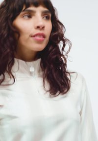 Kvinna med vågigt mörkbrunt hår som bär en vit tröja med dragkedja, tittar något åt sidan mot en enkel vit bakgrund.