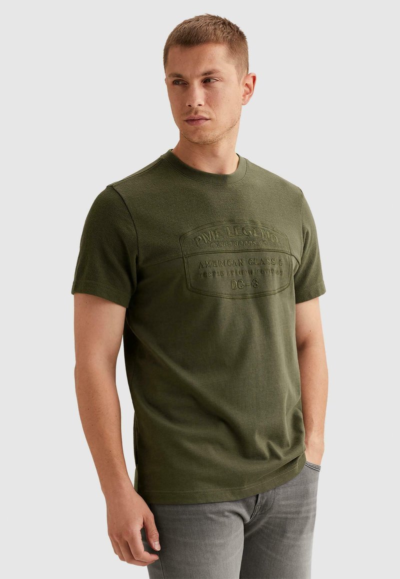 Homme portant un t-shirt à manches courtes vert olive avec texte en relief et un jean gris clair, debout avec une main dans la poche.