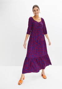 Abito maxi blu con un motivo floreale rosso, con scollo arricciato, maniche a tre quarti e orlo arricciato; indossato con tacchi arancioni.