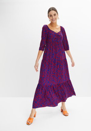 Robe maxi bleue avec un motif floral rouge, présentant un corsage froncé, des manches trois-quarts et un ourlet froncé ; portée avec des talons orange.