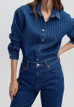 Kvinde iført en blå skjorte med knapper og striber, stukket ned i højtaljede mørkeblå jeans, med den ene hånd på hoften og den anden afslappet.