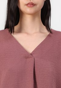 Top color rosa con scollo a V, tessuto strutturato e una sottile piega frontale. Il materiale sembra leggero e si drappeggia morbidamente.