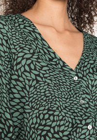 Chemise en tissu noir avec un motif de feuilles vertes, encolure en V et détails de boutons à l'avant, mettant en valeur sa texture légère.