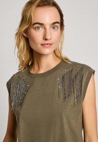 Alyvuogių žalia be rankovių palaidinė su karoliukų ir pūkelių dekoracijomis aplink pečius, pasižyminti lygiu audinio paviršiumi.