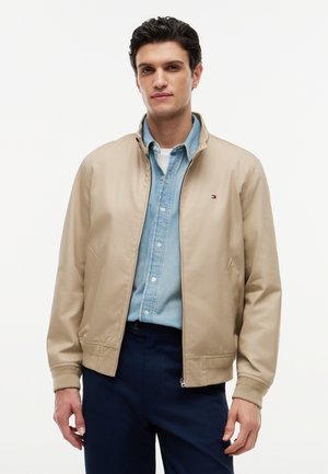 Młody mężczyzna w beżowej kurtce na zamek błyskawiczny, na jasnoniebieskiej koszuli zapinanej na guziki i ciemnoniebieskich spodniach, stoi na tle jednolitego tła.