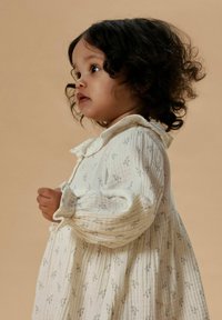 Bambino piccolo con capelli ricci scuri che indossa un vestito color crema a maniche lunghe con piccole stampe floreali, che guarda leggermente verso l'alto su uno sfondo beige.