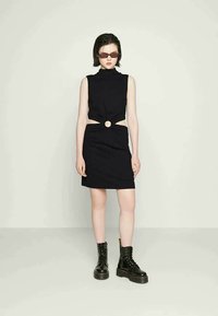 Vestido preto sem mangas com um decote elevado, recortes laterais e um detalhe em forma de círculo na cintura. Combinado com botas de combate pretas.