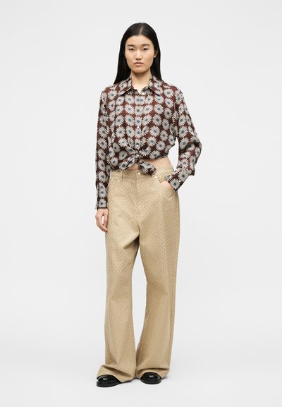 Γυναίκα με μακριά σκούρα μαλλιά που φοράει ένα δεμένο καφέ πουκάμισο με σχέδιο από μαργαρίτες, φαρδιά μπεζ παντελόνια με πουά μοτίβο και μαύρα παπούτσια, στέκεται με το ένα χέρι στην τσέπη.