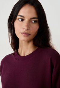 Burgundy sweatshirt med rund halsringning, ribbade muddar och slät yta, bärs av en modell med långt mörkt hår mot en neutral bakgrund.