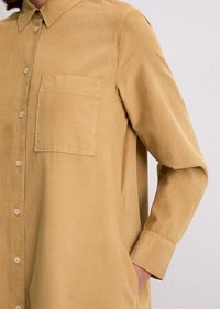 Persoon in een okerkleurig corduroy overhemd met knopen en een borstzak, met één hand in de zijzak, tegen een witte muur als achtergrond.