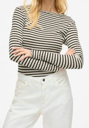 Femme aux bras croisés portant un haut à manches longues rayé noir et blanc et un pantalon blanc taille haute sur fond blanc.
