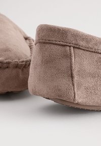 Braune Mokassin-Hausschuhe aus Wildleder mit weicher Textur, rundlicher Spitze und dekorativer Nahtverzierung entlang der Nähte. Flache Gummisohle.