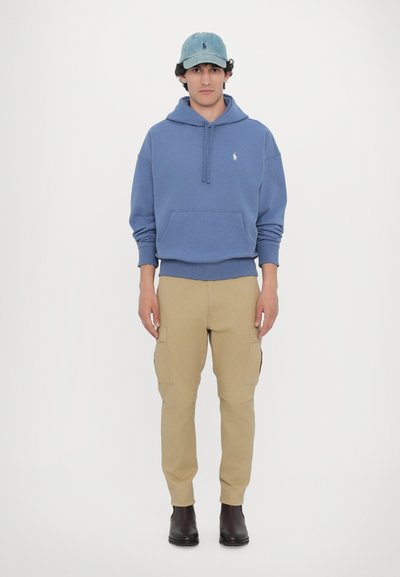 Blå bomulds-hoodie med kængurulomme, tan cargo-bukser og sorte ankelstøvler. Lys blå denimcap med logo-akcenter.