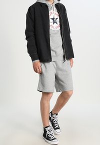 Veste en jean noire sur un sweat à capuche gris avec un logo, associée à un short gris et des baskets montantes noires. Tenue décontractée en couches.